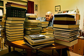 A photo of an older gentleman sitting behind a pile of books.